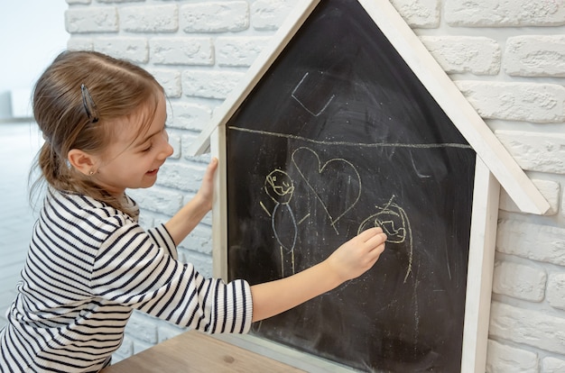 Une petite fille dessine un dessin à la craie sur un tableau noir en forme de maison.