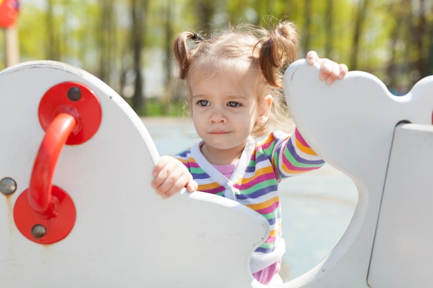 Une petite fille avec deux queues est vêtue d&#39;une veste colorée rayée joue sur le terrain de jeu