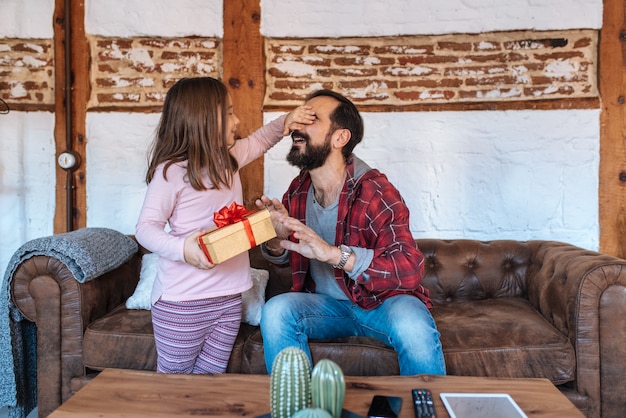 Petite fille donnant un cadeau surprise o son père sur le canapé à la maison le jour de la fête des pères