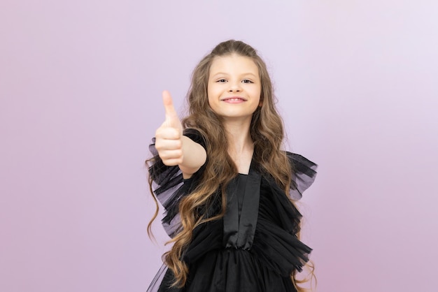 La petite fille donne un coup de pouce et sourit Une mademoiselle aux cheveux ondulés dans une robe noire sur fond rose isolé