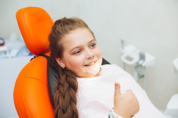 Une petite fille donne un coup de pouce et sourit tout en étant assise dans la médecine et les soins de santé d'une chaise de dentiste