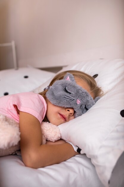 Une petite fille dort dans un masque de sommeil avec une peluche sur le lit dans la chambre