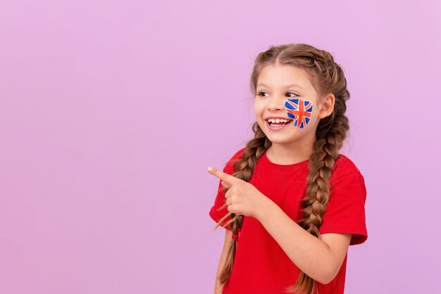 Une petite fille avec un drapeau anglais peint sur sa joue montre le côté et sourit.