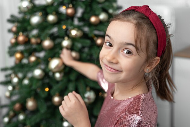 La petite fille drôle accroche des boules de Noël sur l'arbre de Noël, le concept de se préparer pour les vacances de Noël.