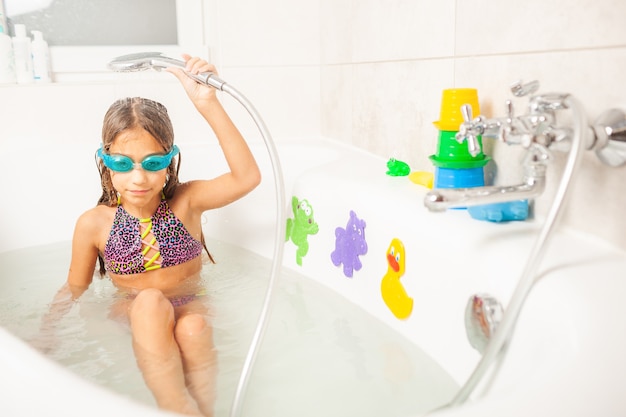 Petite fille drôle dans des verres de bain bleus sourit avec charme tout en versant de l'eau de la douche sur elle-même