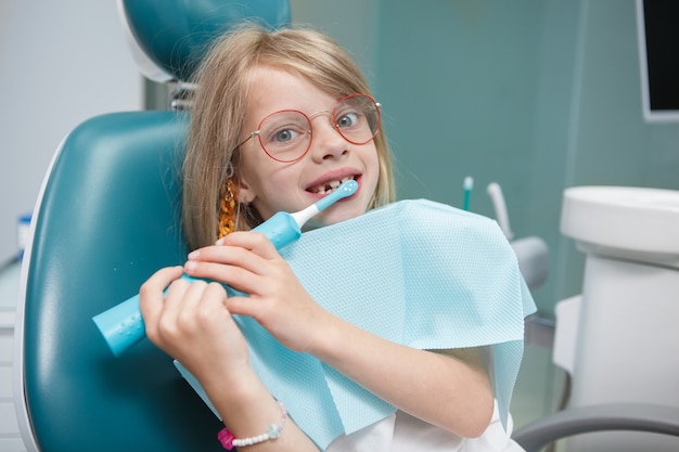 Petite fille drôle regardant la caméra tout en se brossant les dents avec une brosse à dents électrique au bureau du dentiste