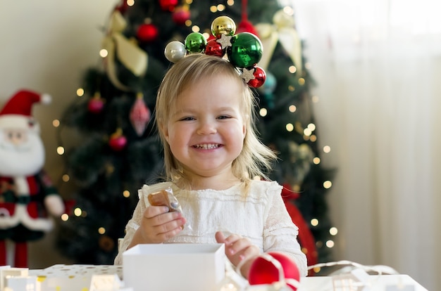 Petite fille drôle en robe blanche ouvrant la boîte-cadeau de Noël à la maison près de l'arbre de Noël