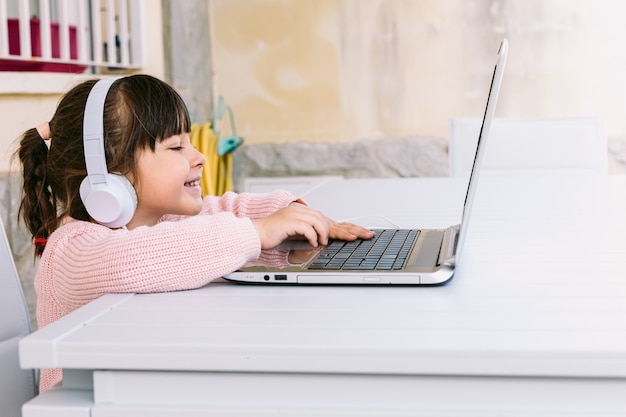Petite fille avec des écouteurs blancs, vêtue d'un pull rose, assise devant un ordinateur portable, tapant, sur la terrasse, souriante. Concept d'étude à distance en ligne, enfance et technologie.