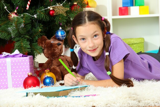 Petite fille écrivant une lettre au Père Noël près de l'arbre de Noël