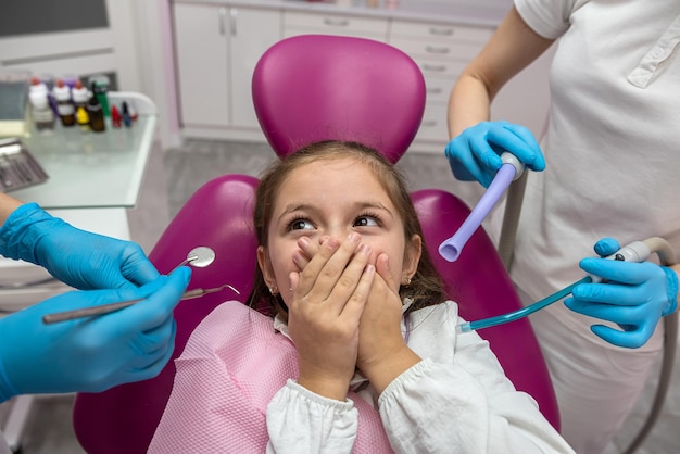 Petite fille effrayée couvrant sa bouche avec ses mains lors d'une visite chez le dentiste Gros plan d'un orthodontiste essayant de vérifier les dents d'un enfant effrayé assis dans un fauteuil dentaire dans une clinique