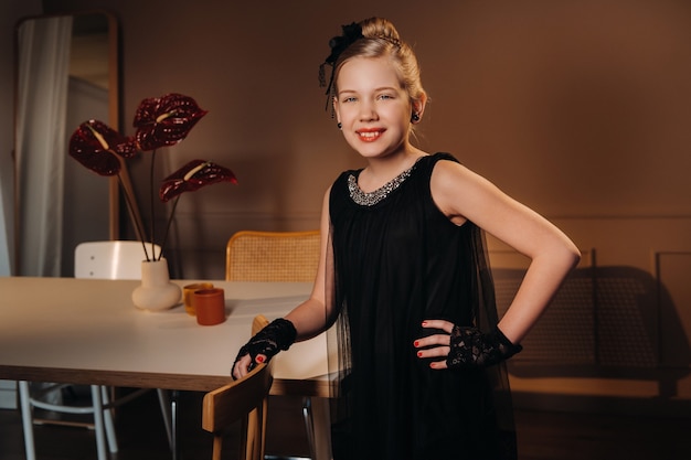 Une petite fille élégante dans une robe noire se tient à l'intérieur près de la table.