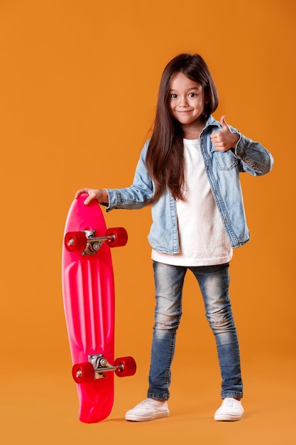 Petite fille élégante avec une planche à roulettes en vêtements de jeans sur fond orange