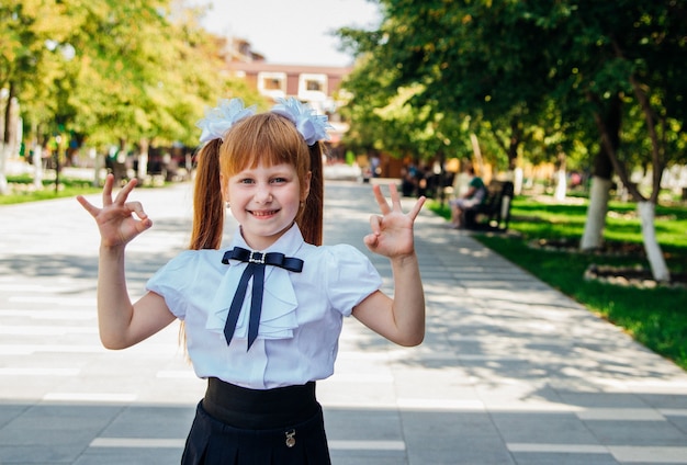Une petite fille, une élève du primaire, se tient dans la rue, une écolière montre un geste d'approbation avec ses mains OK. Portrait d'une écolière aux cheveux rouges. Retour à l'école.