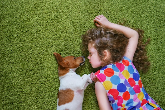 Petite fille embrassant le chiot jack russell et se trouvant sur le tapis vert. Haute vue de dessus.