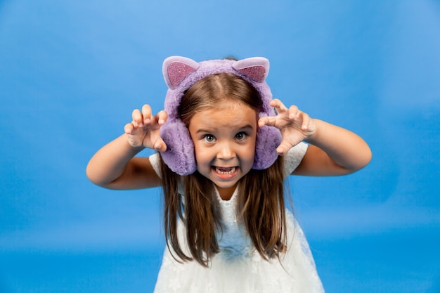 Petite fille émotionnelle en colère dans un casque de fourrure sur fond bleu.