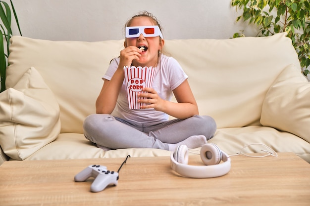 Petite fille émotive dans des lunettes de film 3d mangeant du pop-corn dans un seau rayé et regardant la télévision à la maison