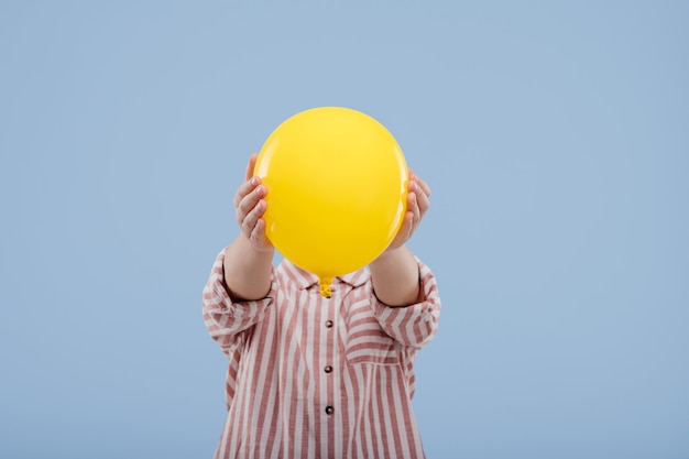 Petite fille enfant couvrant le visage avec un ballon jaune, vêtue d'une chemise rayée, isolée sur fond bleu, espace pour copie