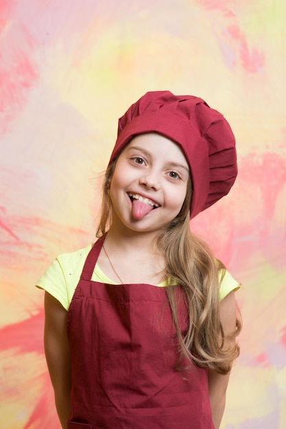 Petite fille ou enfant mignon avec un visage heureux montrant la langue en toque rouge et tablier de cuisine sur fond abstrait coloré
