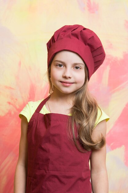 Petite fille ou enfant mignon avec un visage heureux en toque rouge et tablier de cuisinier sur fond abstrait coloré