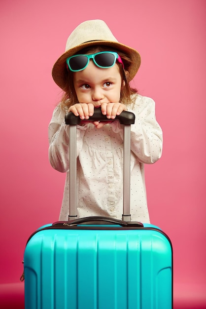 Une petite fille ennuyée avec une valise bleue porte un chapeau de paille et des lunettes de soleil exprime sa perplexité