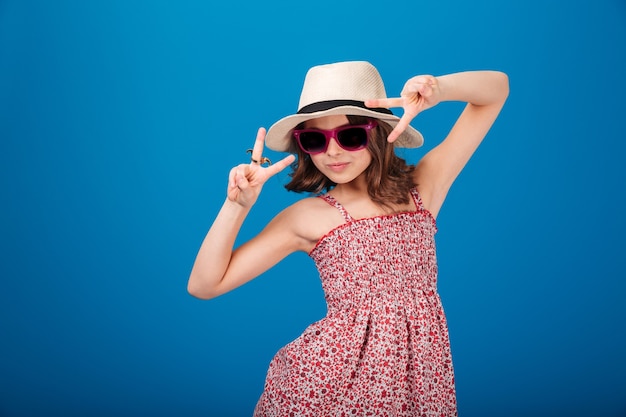 Petite fille espiègle mignonne dans le chapeau, la robe d'été et les lunettes de soleil montrant le signe de paix avec les deux mains au-dessus du fond bleu
