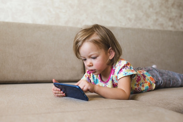 Une petite fille est allongée sur le canapé et regarde le téléphone