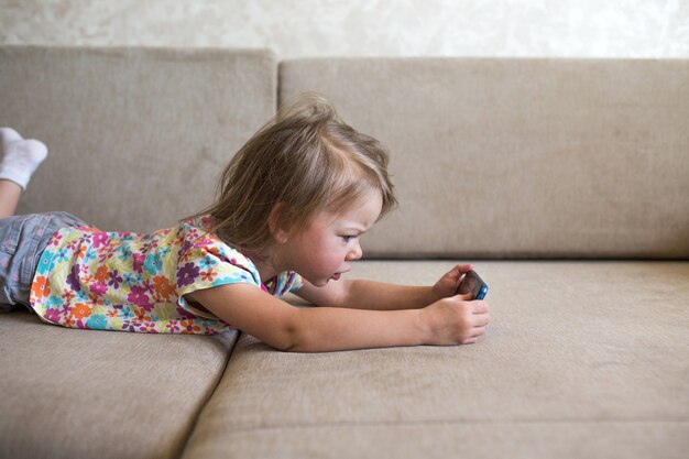 Une petite fille est allongée sur le canapé et regarde le téléphone