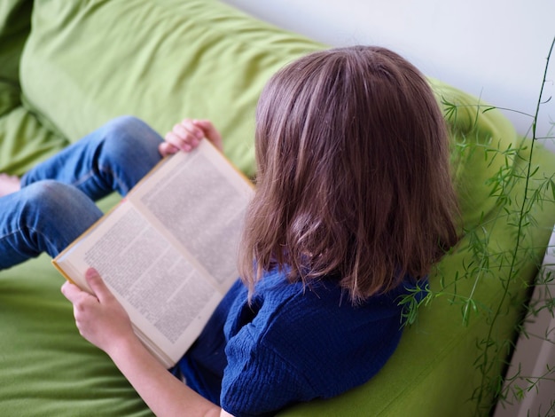 une petite fille est allongée sur le canapé vert et lit un livre à la maison