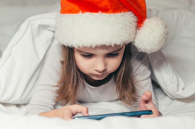 Une petite fille est allongée dans son lit avec un chapeau de père Noël et regarde des dessins animés, étudie, joue à un jeu au téléphone