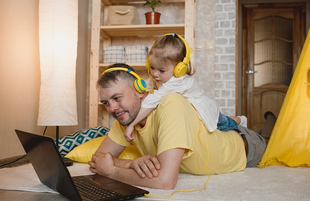 Une petite fille est allongée sur le dos de son père et écoute de la musique avec des écouteurs jaunes tout en regardant son ordinateur portable. le concept de bonheur familial