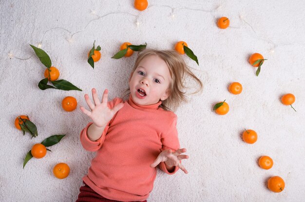 Une petite fille est allongée sur le sol clair, beaucoup de mandarines sont éparpillées autour d'elle.