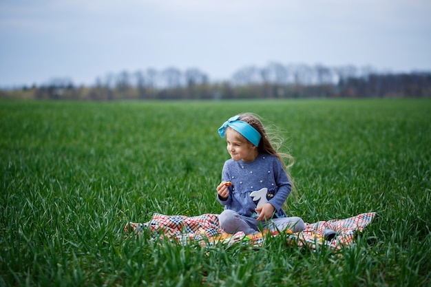 Une petite fille est assise sur le couvre-lit et mange des biscuits et de la marmelade, de l'herbe verte sur le terrain, un printemps ensoleillé, le sourire et la joie de l'enfant, un ciel bleu avec des nuages