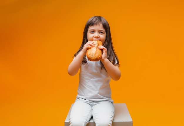 Une petite fille est assise sur un cube sur un fond jaune et mange un pain des aliments sains des produits naturels du pain