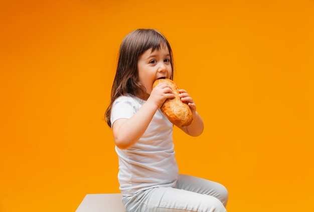 Une petite fille est assise sur un cube sur un fond jaune et mange un pain des aliments sains des produits naturels du pain