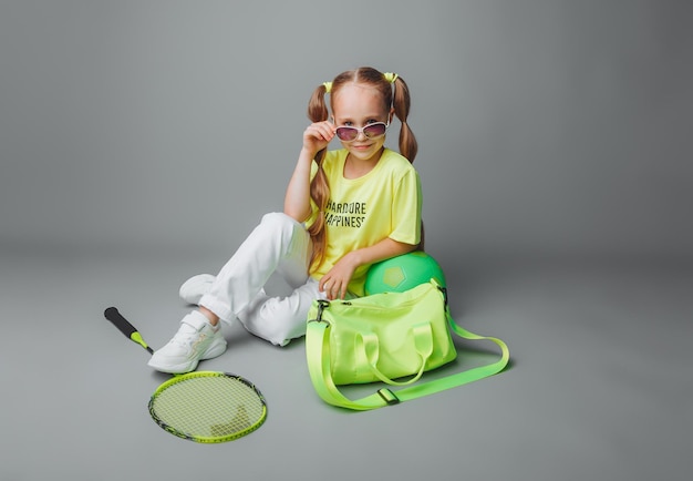 Une petite fille est assise sur un fond gris avec une raquette une balle et un sac de sport un petit athlète sports pour enfants contenu sportif