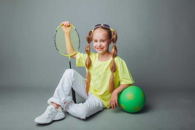 Une petite fille est assise sur un fond gris avec une raquette une balle et un sac de sport un petit athlète sports pour enfants contenu sportif