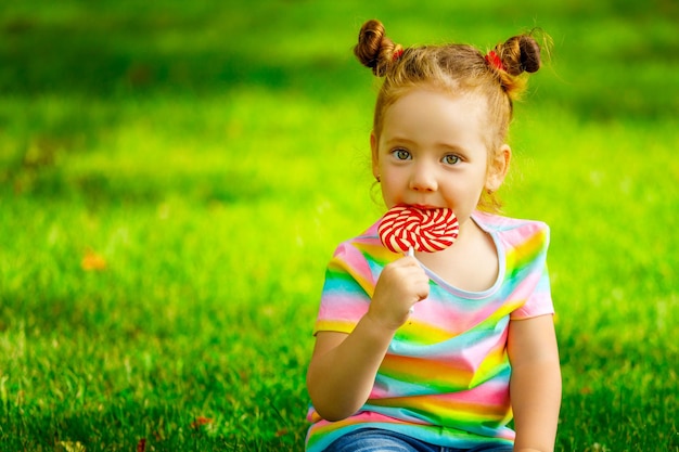 Une petite fille est assise sur l'herbe dans le parc et met une sucette dans sa bouche