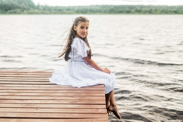 Une petite fille est assise sur une jetée près du lac de la ville à l'extérieur. vacances d'été