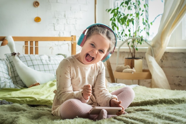 Une petite fille est assise sur le lit dans un casque bébé joue imprudemment à des jeux et écoute de la musique