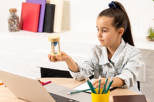 petite fille est assise avec un ordinateur portable à table et tient un sablier, attendant la fin des cours en ligne