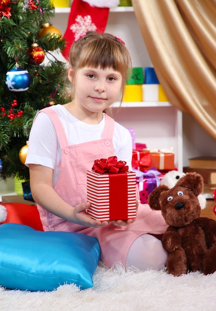 Une petite fille est assise près d'un arbre de Noël avec un cadeau à la main dans une pièce décorée de façon festive