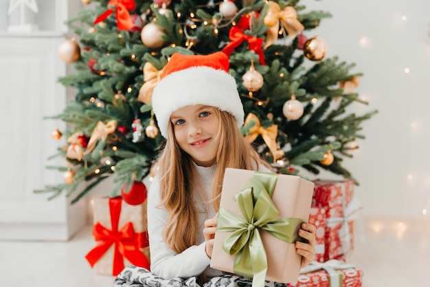 Une petite fille est assise près du sapin de Noël avec un cadeau de Noël. Joyeux Noël.