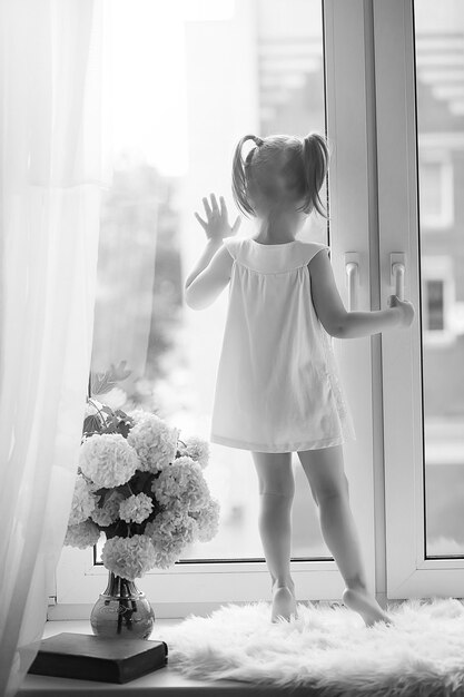 Une petite fille est assise sur le rebord de la fenêtre. Un bouquet de fleurs dans un vase près de la fenêtre et une fille reniflant des fleurs. Une petite princesse en robe blanche avec un bouquet de fleurs blanches par fenêtre.