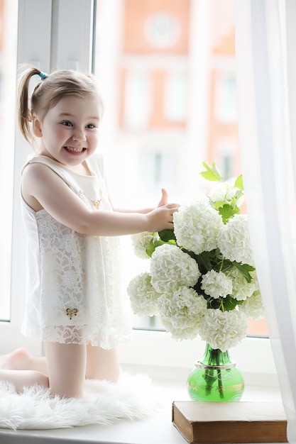 Une petite fille est assise sur le rebord de la fenêtre. Un bouquet de fleurs dans un vase près de la fenêtre et une fille reniflant des fleurs. Une petite princesse en robe blanche avec un bouquet de fleurs blanches près de la fenêtre.