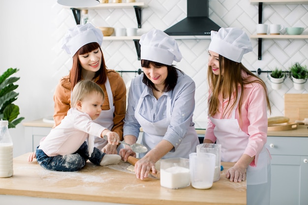 Petite fille est assise sur la table en bois de la cuisine pendant que sa mère, sa tante et sa grand-mère font la pâte à biscuits. Femmes heureuses en tabliers blancs cuire ensemble