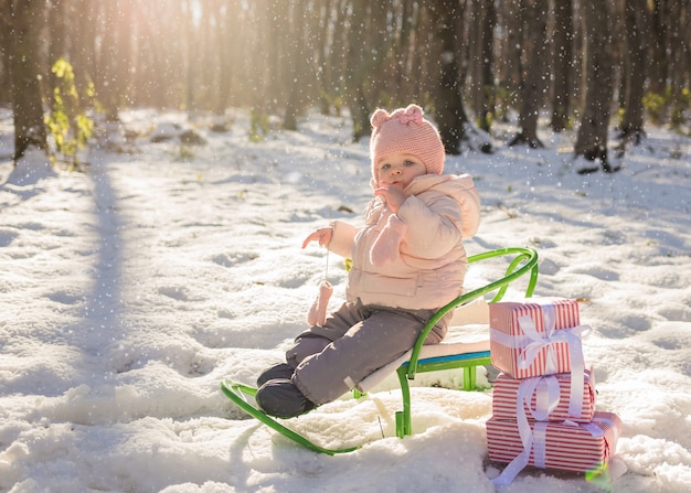 Une petite fille est assise sur un traîneau dans une forêt d'hiver avec des cadeaux de chutes de neige