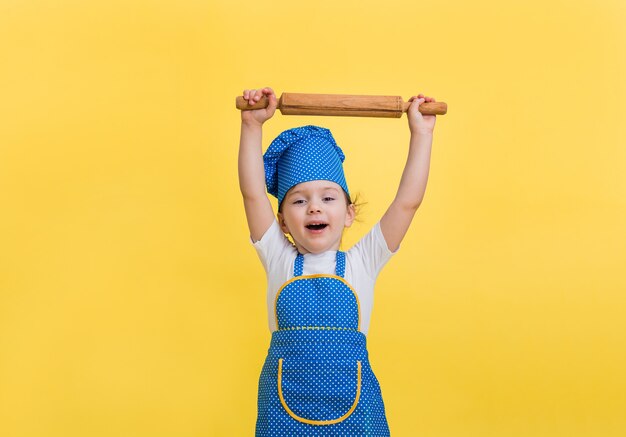 Une petite fille est heureuse dans un tablier de cuisine et un chapeau avec un rouleau à pâtisserie sur un espace jaune. Vous cherchez.