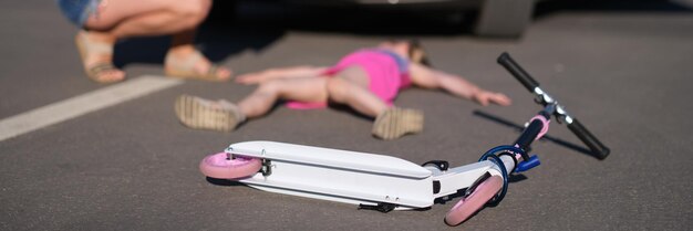 Une petite fille est immobile sur l'asphalte avec un scooter après une collision avec une voiture