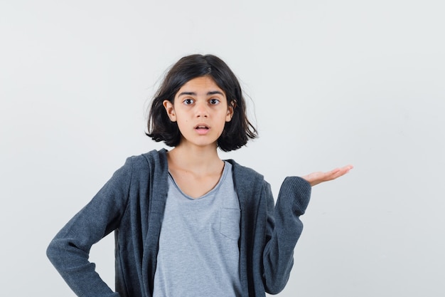 Petite fille étalant la paume de côté en t-shirt, veste et à la perplexité, vue de face.