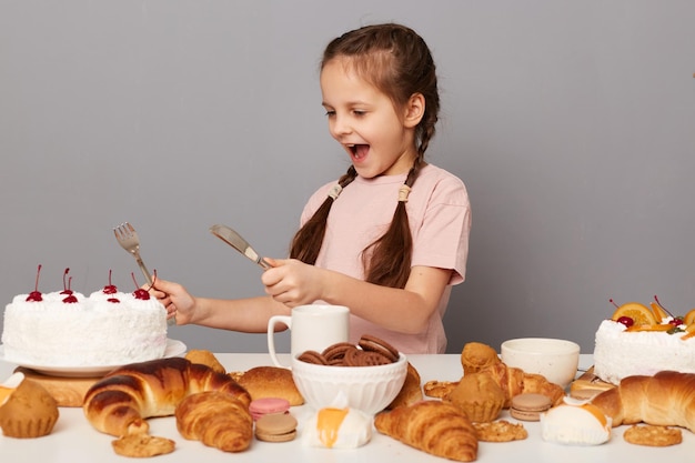 Une petite fille étonnée et excitée avec des nattes assis à table isolée sur fond gris tenant une fourchette et un couteau a l'air affamée de manger un gros gâteau savoureux